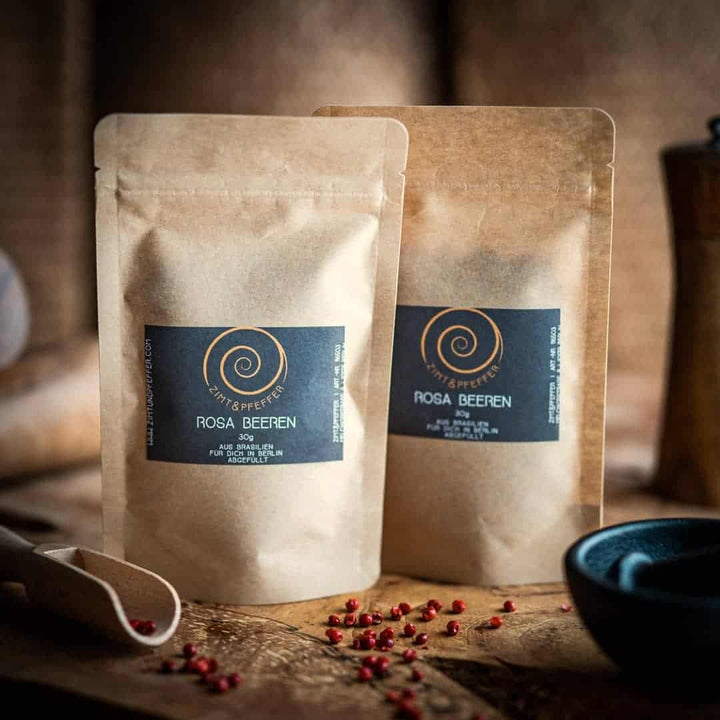 Zwei Tüten aus Kraftpapier stehen auf einem Olivenholzbrett. Die Zip-Tüten sind mit Rosa Beeren gefüllt und haben jeweils eine Füllmenge von 30g.