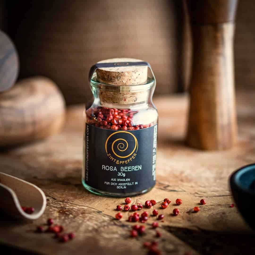 Ein Glas mit Presskorken steht auf einem Olivenholzbrett. Das Glas ist mit Rosa Beeren gefüllt und hat eine Füllmenge von 30g.