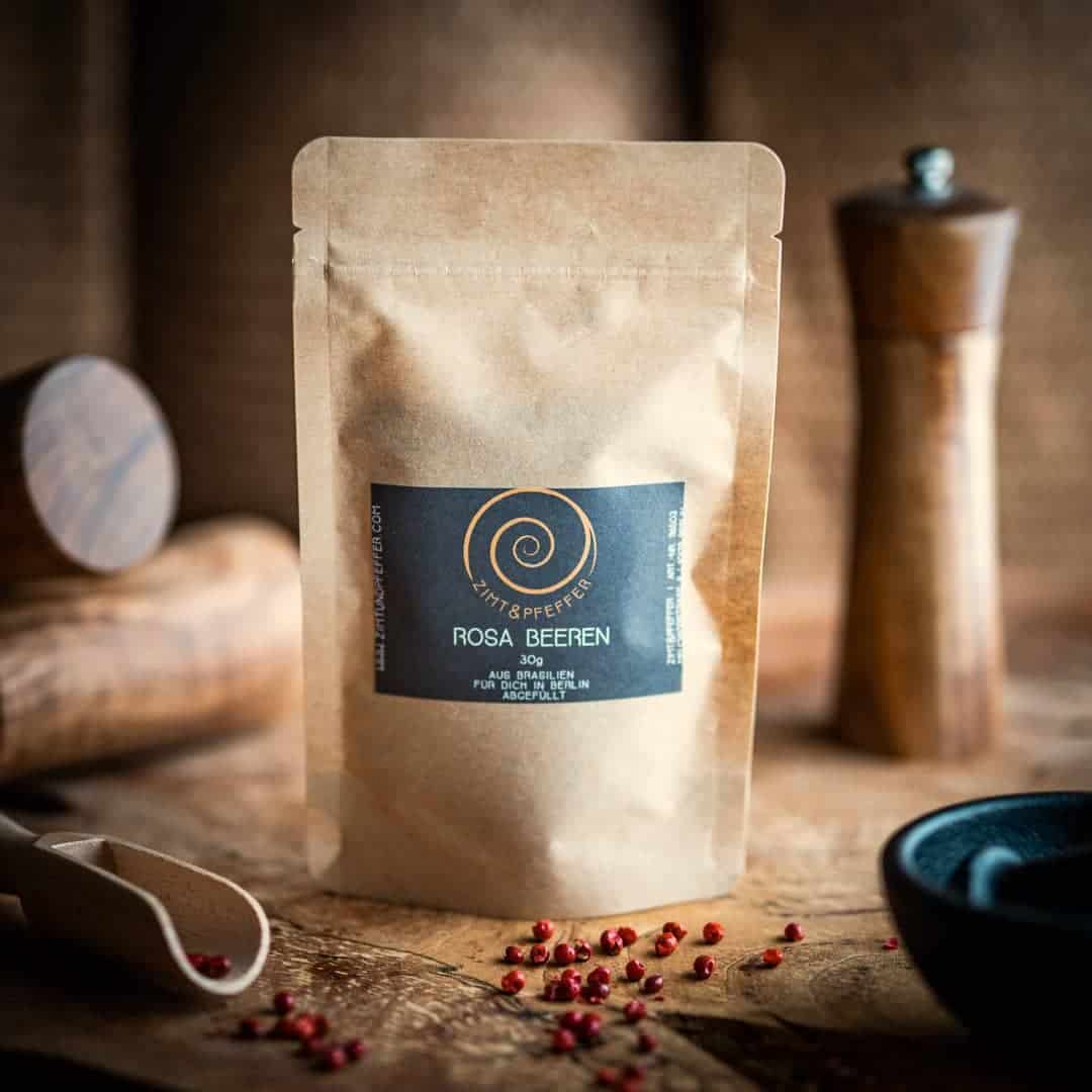 Eine Tüte aus Kraftpapier steht auf einem Olivenholzbrett. Die Zip-Tüte ist mit Rosa Beeren gefüllt und hat eine Füllmenge von 30g.