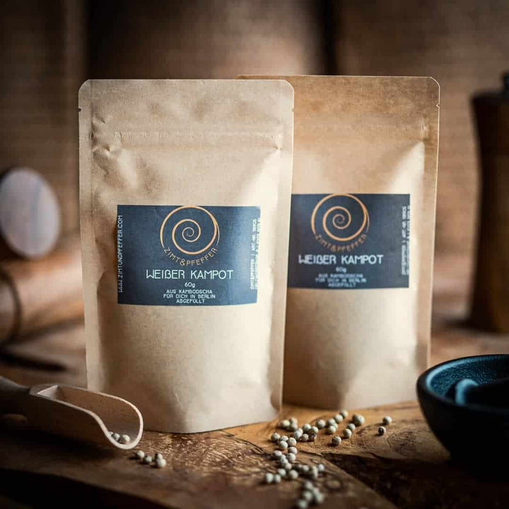 Zwei Tüten aus Kraftpapier stehen auf einem Olivenholzbrett. Die Zip-Tüten sind mit Weißer Kampot Pfeffer gefüllt und haben jeweils eine Füllmenge von 60g.