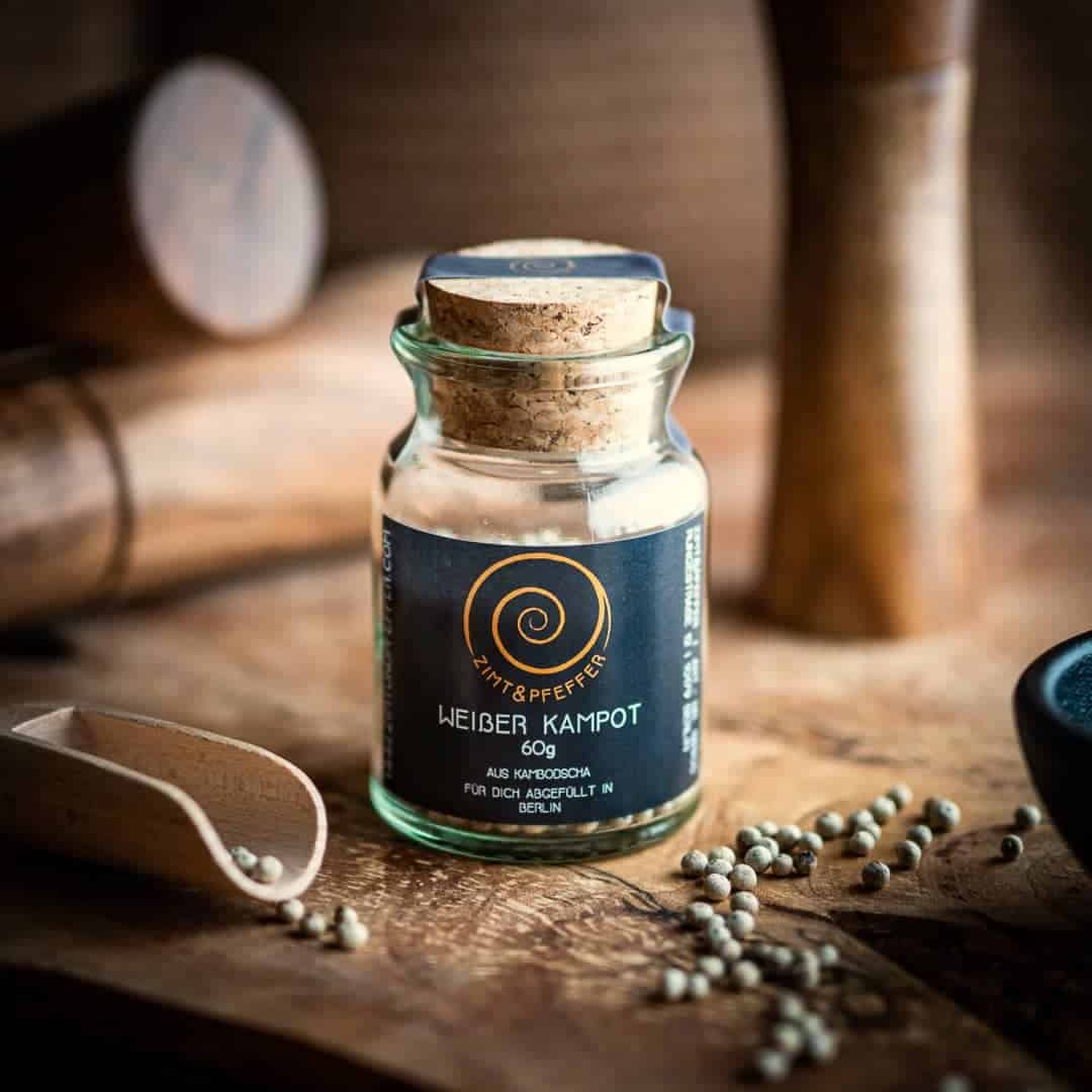Ein Glas mit Presskorken steht auf einem Olivenholzbrett. Das Glas ist mit Weißer Kampot Pfeffer gefüllt und hat eine Füllmenge von 60g.
