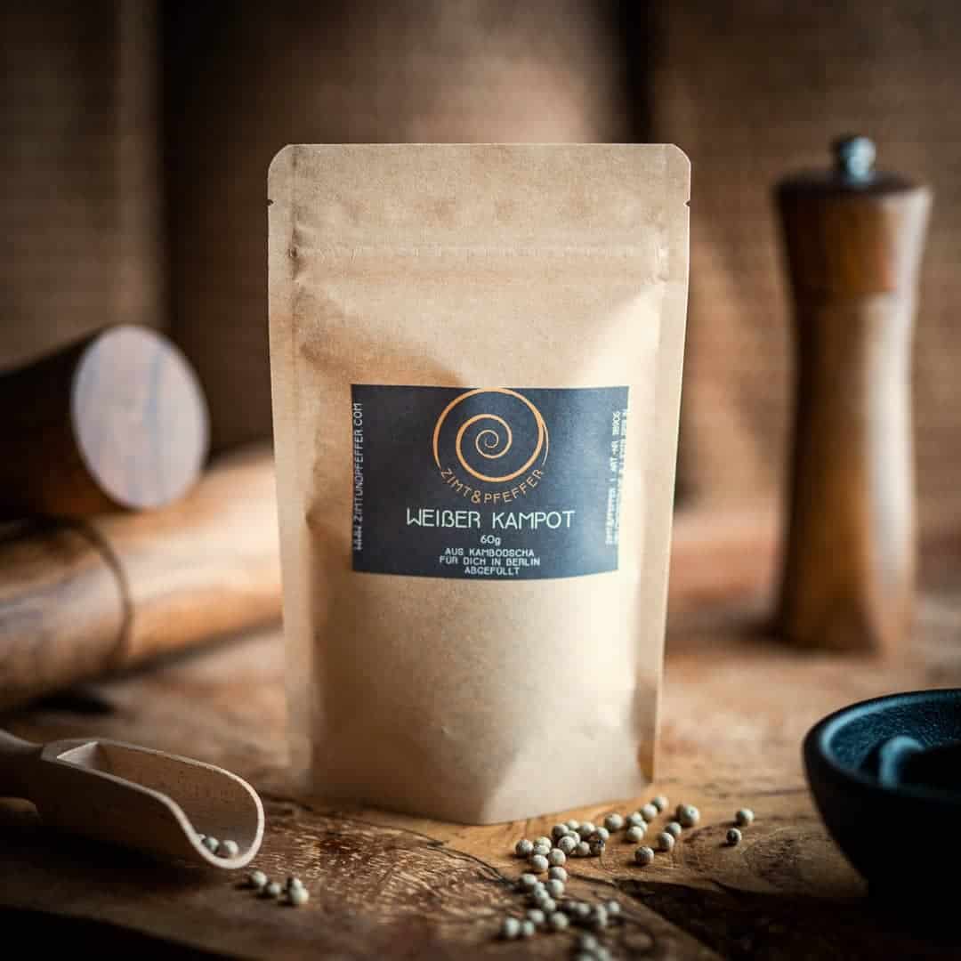 Eine Tüte aus Kraftpapier steht auf einem Olivenholzbrett. Die Zip-Tüte ist mit Weißer Kampot Pfeffer gefüllt und hat eine Füllmenge von 60g.