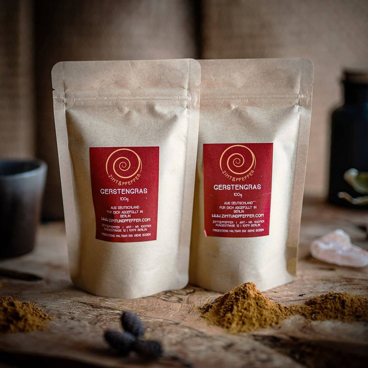 Zwei 80g Tüten Gerstengras Pulver auf einem Olivenholzbrett.