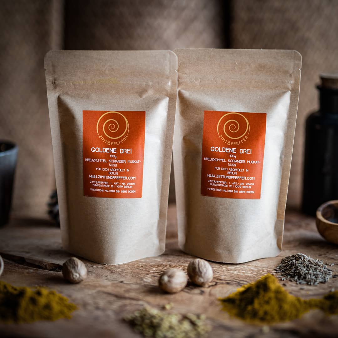 Zwei 100g Tüten Goldene Drei auf einem Olivenholzbrett. Bei dem Produkt handelt es sich um eine Gewürzmischung aus Kreuzkümmel, Koriander und Muskat.
