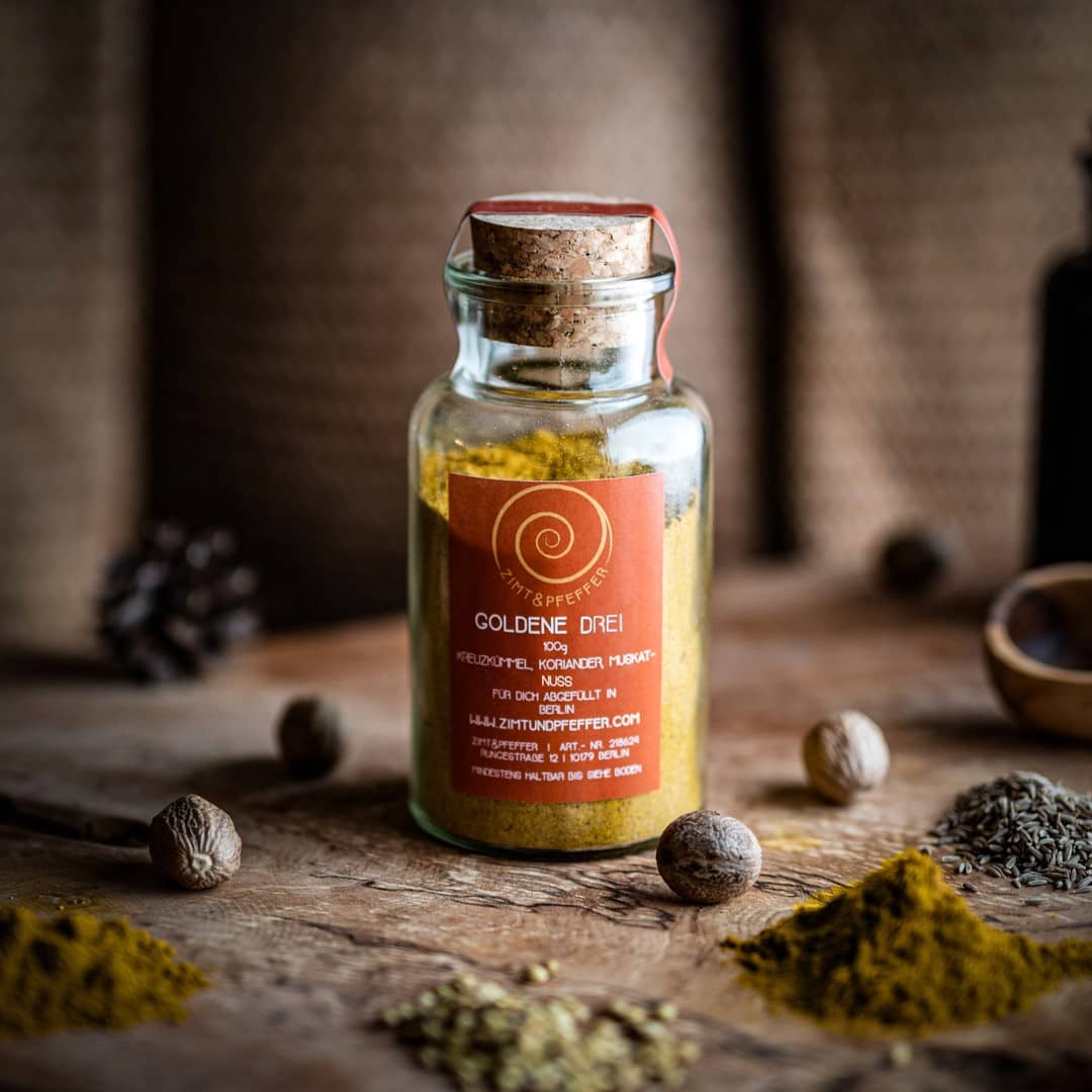 Ein 100g Glas Goldene Drei auf einem Olivenholzbrett. Bei dem Produkt handelt es sich um eine Gewürzmischung aus Kreuzkümmel, Koriander und Muskat.