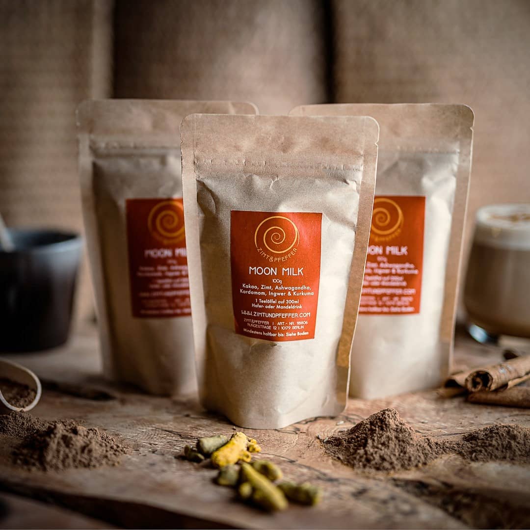 Drei 100g Tüten Moon Milk auf einem Olivenholzbrett. Bei dem Produkt handelt es sich um eine Mischung aus purem Rohkakao, Zimt, Ashwagandha, Kardamom, Ingwer und Kurkuma.