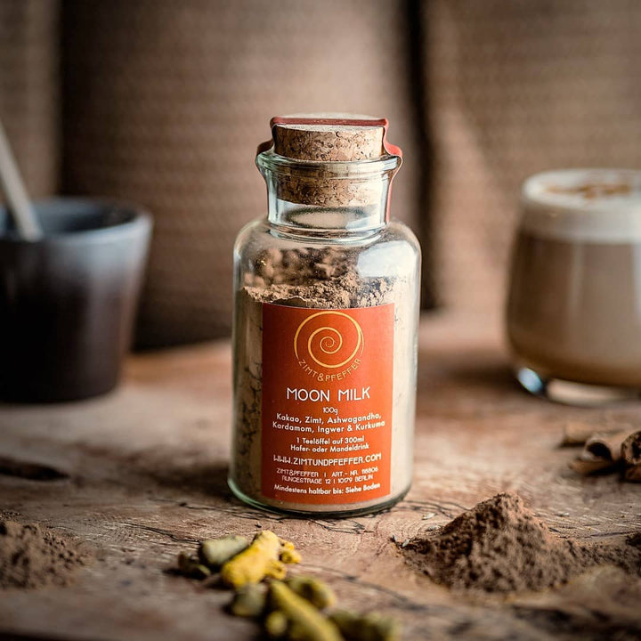 Ein 100g Glas Moon Milk auf einem Olivenholzbrett. Bei dem Produkt handelt es sich um die Mischung Moon Milk, aus purem Rohkakao, Zimt, Ashwagandha, Kardamom, Ingwer und Kurkuma.