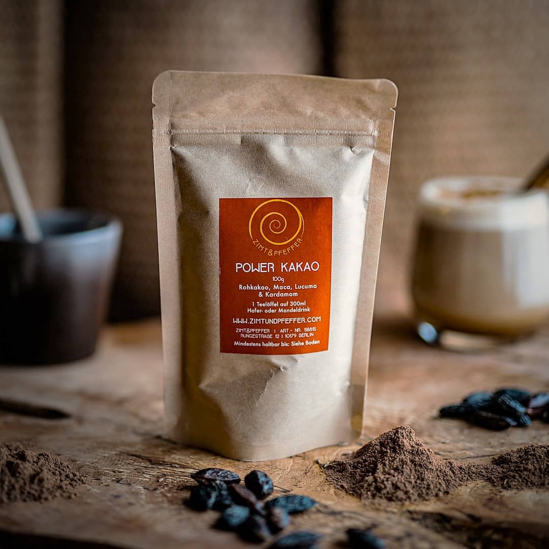 Eine 100g Tüte Power Kakao auf einem Olivenholzbrett. Eine Mischung aus Rohkakao, Maca, Kardamom und Lucuma.