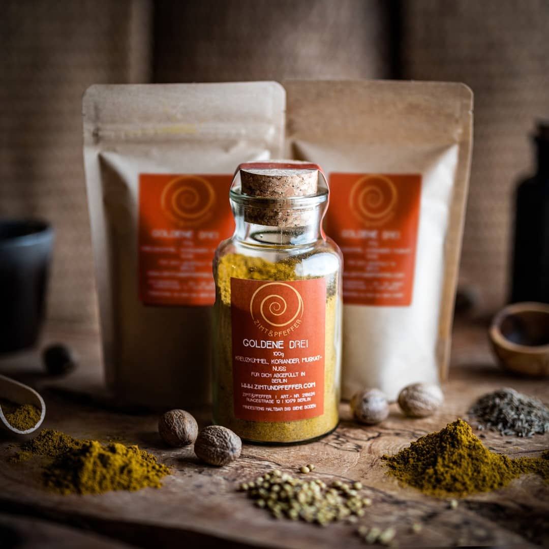 Ein 100g Glas und 2 100g Tüten Goldene Drei auf einem Olivenholzbrett. Bei dem Produkt handelt es sich um eine Gewürzmischung aus Kreuzkümmel, Koriander und Muskat.