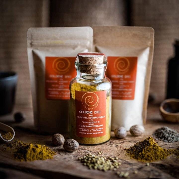 Ein 100g Glas und 2 100g Tüten Goldene Drei auf einem Olivenholzbrett. Bei dem Produkt handelt es sich um eine Gewürzmischung aus Kreuzkümmel, Koriander und Muskat.