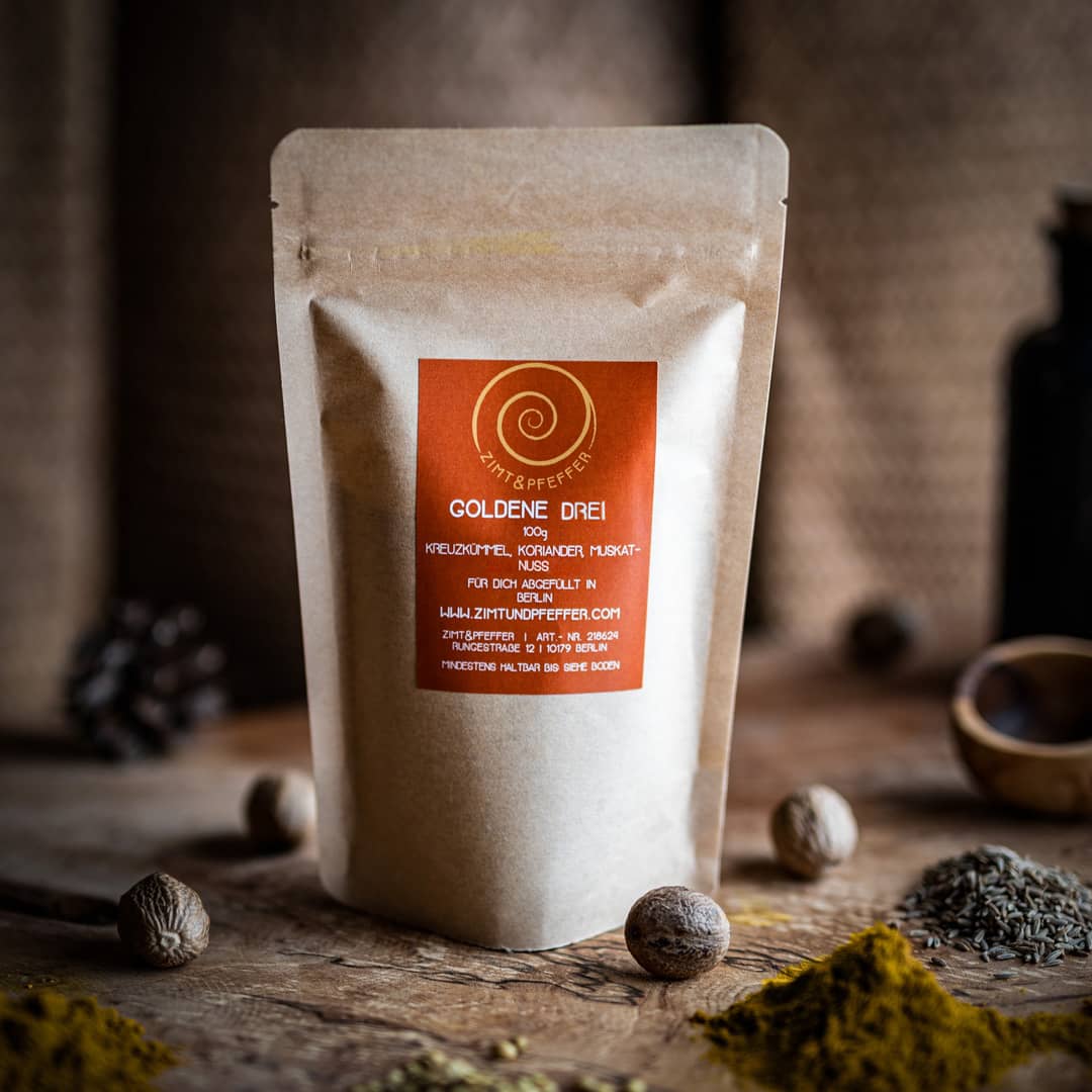 Eine 100g Tüte Goldene Drei auf einem Olivenholzbrett. Bei dem Produkt handelt es sich um eine Gewürzmischung aus Kreuzkümmel, Koriander und Muskat.