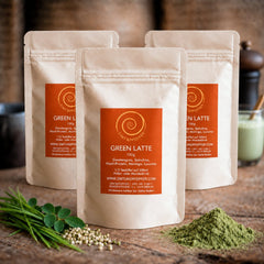 Drei 100g Tüten mit Green Latte auf einem Olivenholzbrett. Eine Mischung aus Gerstengras, Spirulina, Hanf und Lucuma.