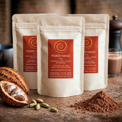 Drei 100 g Tüten Power Kakao auf einem Olivenholzbrett. Eine Mischung aus Rohkakao, Maca, Kardamom und Lucuma.