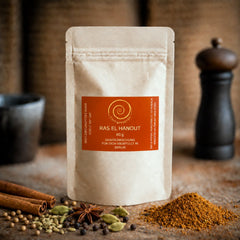 Ein Nachfüllpack Ras el Hanout steht auf einem Olivenholzbrett. Die nordafrikanische Mischung besteht aus Kurkuma, Koriander, Fenchel, Zimt, Pfeffer, Ingwer, Piment, Cumin, Chili, Kardamom, Bockshornkleesaat, Muskatnuss und Anis und hat eine Füllmenge von 60 g.