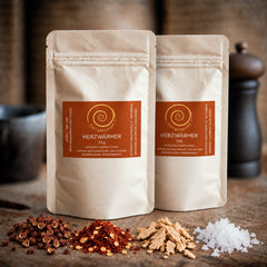 Zwei Tüten Herzwärmer mit je 50 g auf einem Olivenholzbrett. Eine harmonisch abgestimmte Gewürzzubereitung aus Szechuan Pfeffer, Chili- und Ingwerflocken sowie feinstem Pyramidensalz.