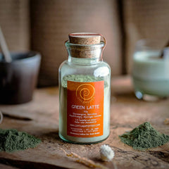 Ein 100g Glas mit Green Latte auf einem Olivenholzbrett. Eine Mischung aus Gerstengras, Spirulina, Hanf, Moringa und Lucuma.