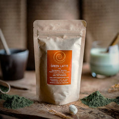 Eine 100g Tüte mit Green Latte auf einem Olivenholzbrett. Eine Mischung aus Gerstengras, Spirulina, Hanf, Moringa und Lucuma.