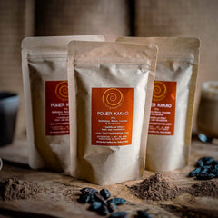 Drei 100g Tüten Power Kakao auf einem Olivenholzbrett. Eine Mischung aus Rohkakao, Maca, Kardamom und Lucuma.