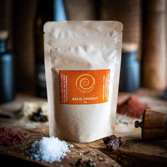 Ein Nachfüllpack Ras el Hanout steht auf einem Olivenholzbrett. Die nordafrikanische Mischung besteht aus Kurkuma, Koriander, Fenchel, Zimt, Pfeffer, Ingwer, Piment, Cumin, Chili, Kardamom, Bockshornkleesaat, Muskatnuss und Anis und hat eine Füllmenge von 60g.