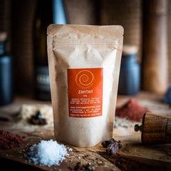 Eine 60g Tüte Zahtar auf einem Olivenholzbrett. Eine Gewürzmischung aus Sesamsaat, Zumaq (Sumachfrüchte gemahlen), Thymian und Oregano.