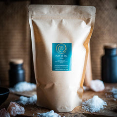 Eine Zip-Tüte aus Kraftpapier steht auf einem Olivenholzbrett. Die Zip-Tüte ist mit Flor de Sal gefüllt und hat eine Füllmenge von 1kg.