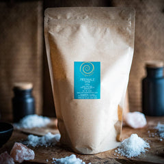 Eine Zip-tüte aus Kraftpapier steht auf einem Oliven Holzbrett vor einem sandfarbenem Hintergrund. Die Zip-Tüte ist mit grobem portugiesischem Meersalz gefüllt und hat eine Füllmenge von 1kg.