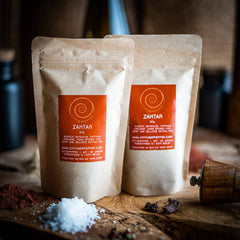 Zwei 60g Tüten Zahtar auf einem Olivenholzbrett. Eine Gewürzmischung aus Sesamsaat, Zumaq (Sumachfrüchte gemahlen), Thymian und Oregano.